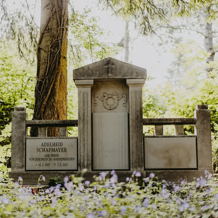 Historisches Grabmal mit steinerner Umrandung und Blumen im Vordergrund auf einem friedlichen Waldfriedhof